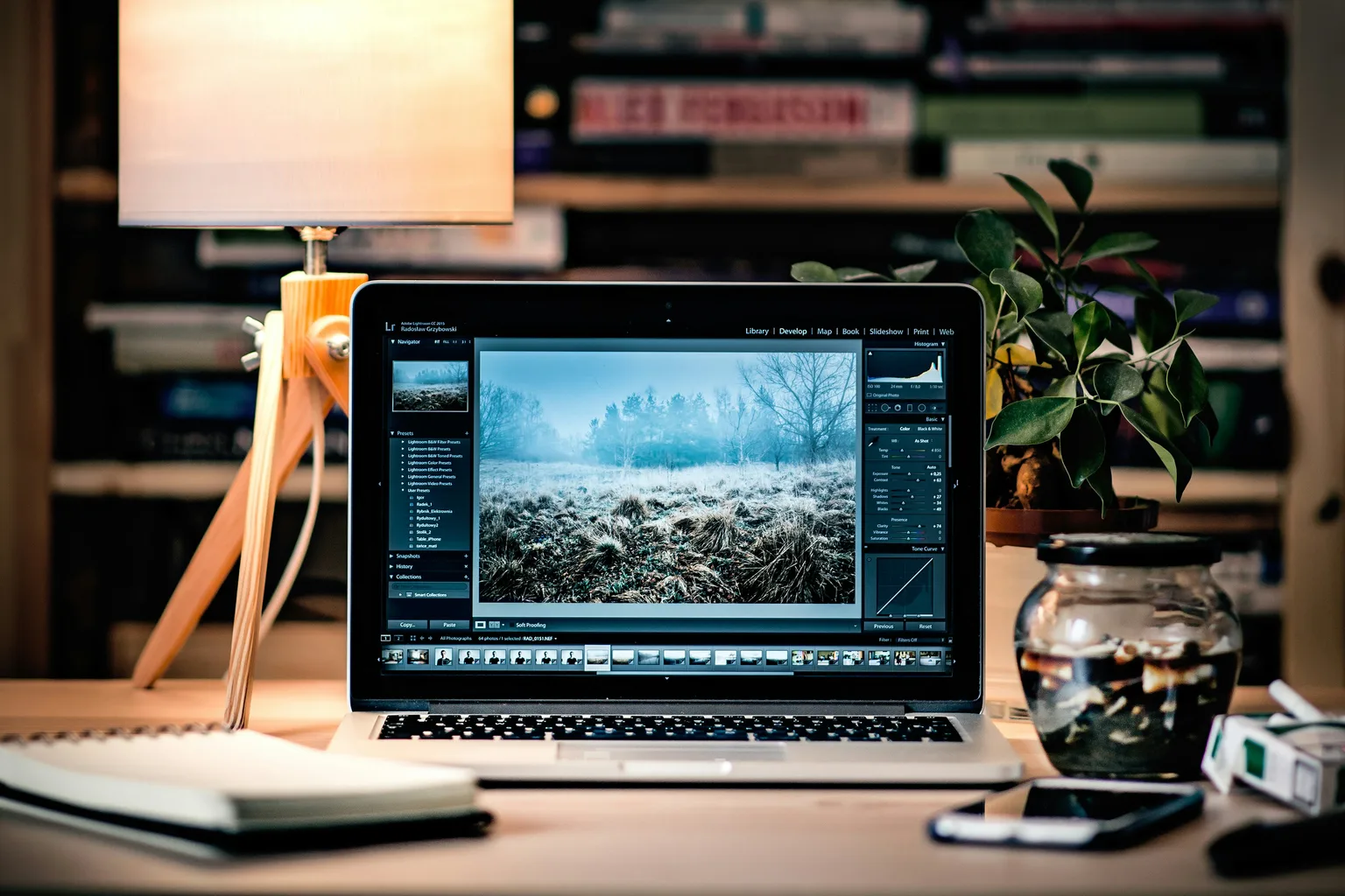 An open MacBook Pro on a wooden desk beside a table lamp, a notebook and a potted plant, with bookshelves behind.